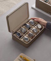A person arranges a Fraser 8 Watch Case - Taupe from TAWBURY that holds eight watches. Nearby, a tool and another case suggest the meticulous world of watch collectors.